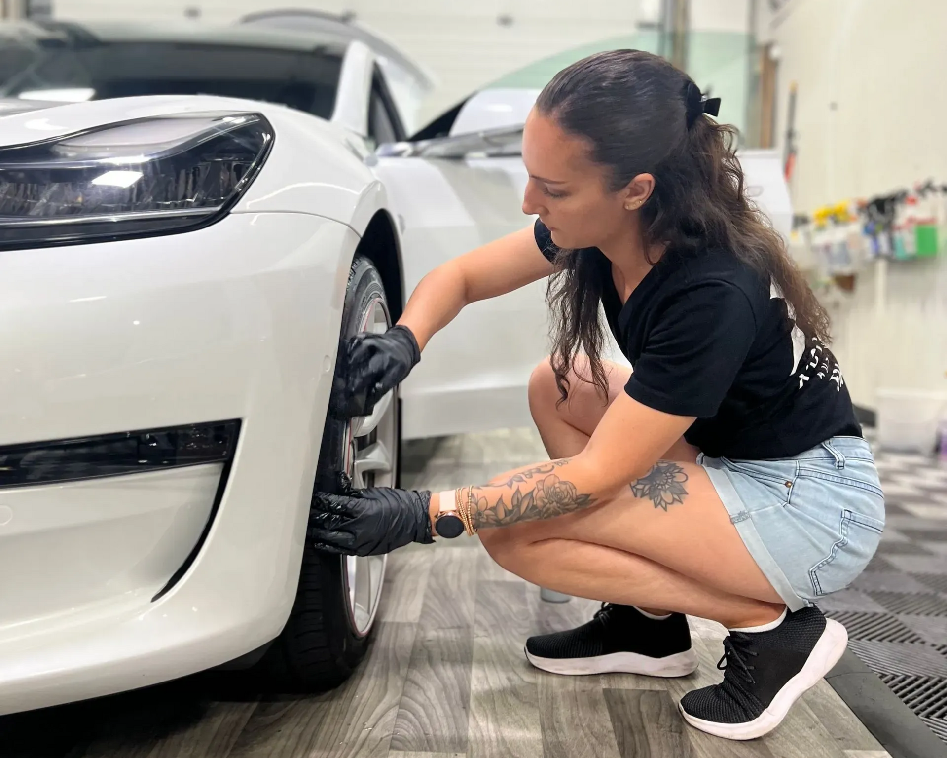 A person wearing black gloves squats to detail the tire of a white car in a garage setting.