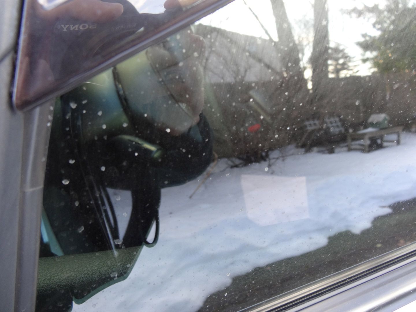 A reflection of a person in a car window with snow on the ground