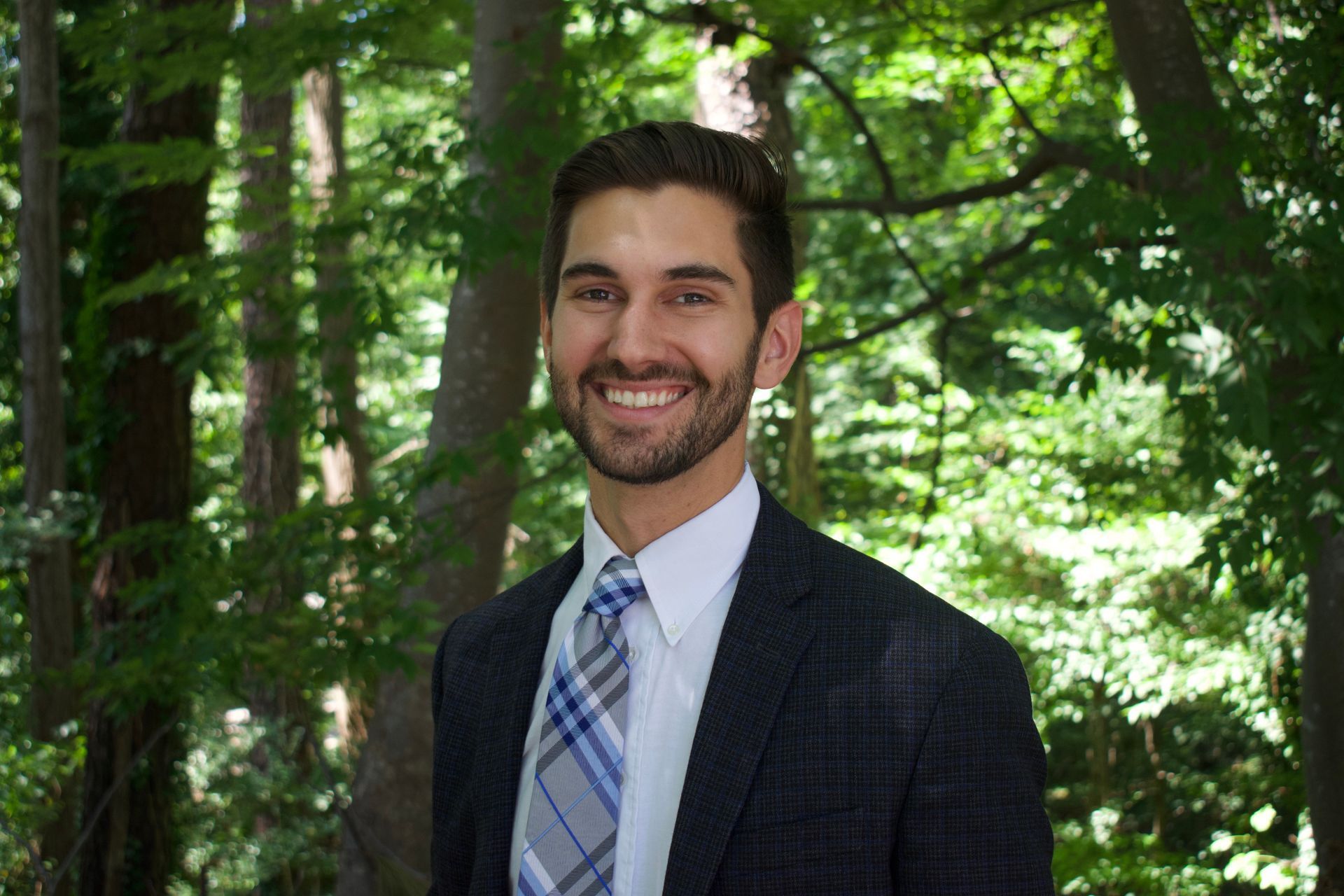 Smiling man in a suit and tie standing in a sunlit forest.