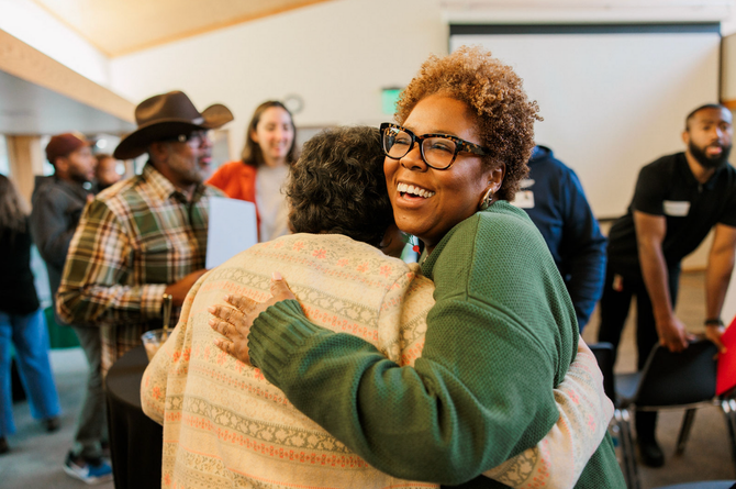Two people hugging in a bright room while others stand and chat in the background.