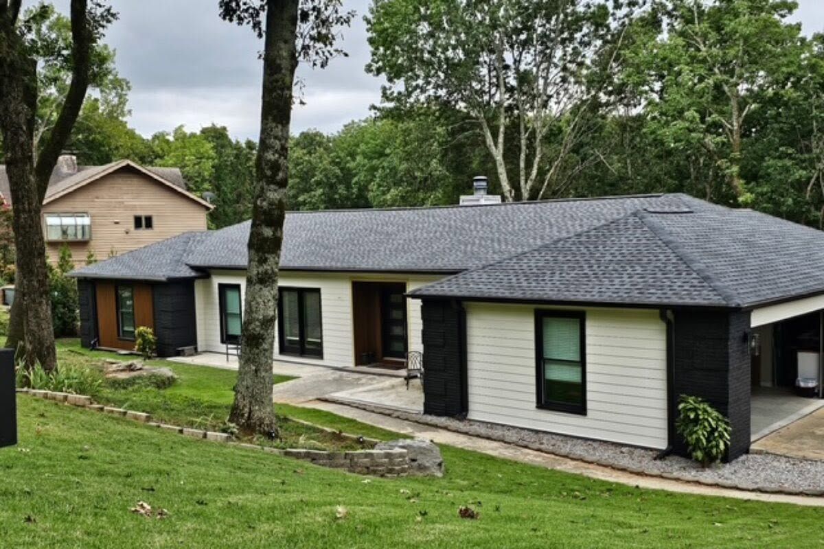 Single-story house with gray roof, white siding, and large windows on a wooded hillside.
