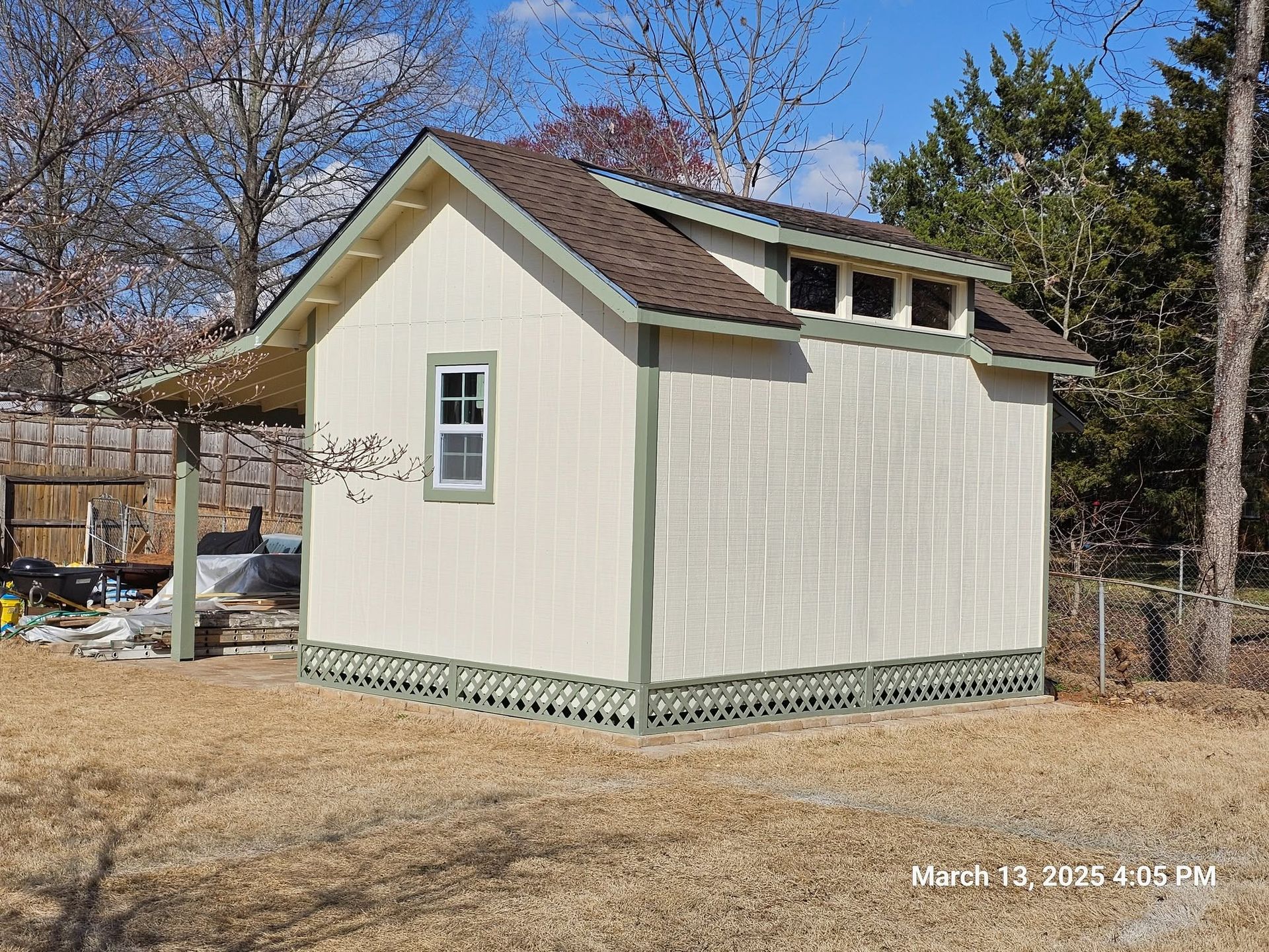 Small beige shed with white trim and a sloped roof in a backyard, trees and fence behind it.
