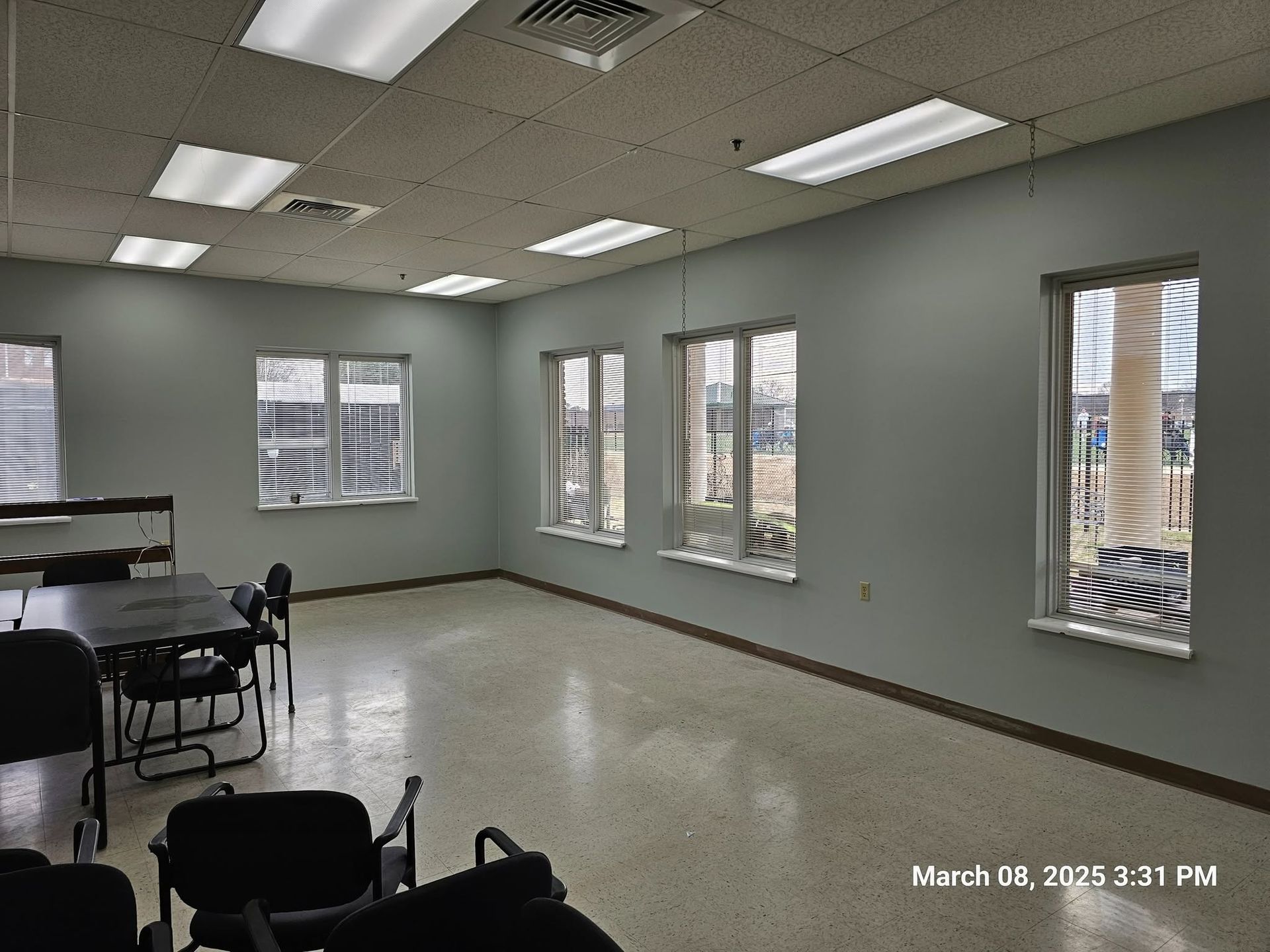 Empty classroom with gray walls, fluorescent lights, tables, chairs, and windows along one side.
