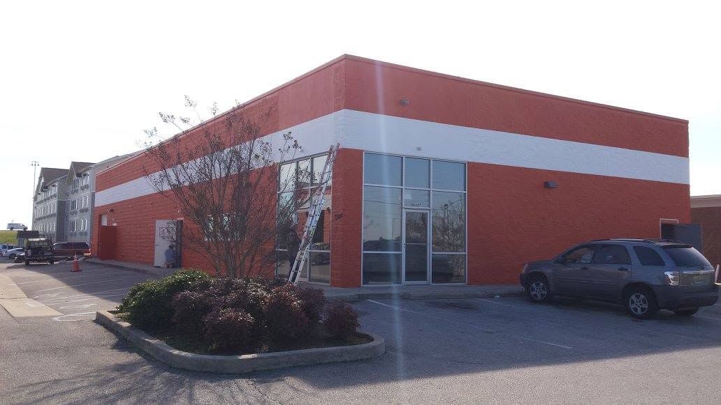 Red and white commercial building with glass entrance, parked SUV, and landscaped shrubs