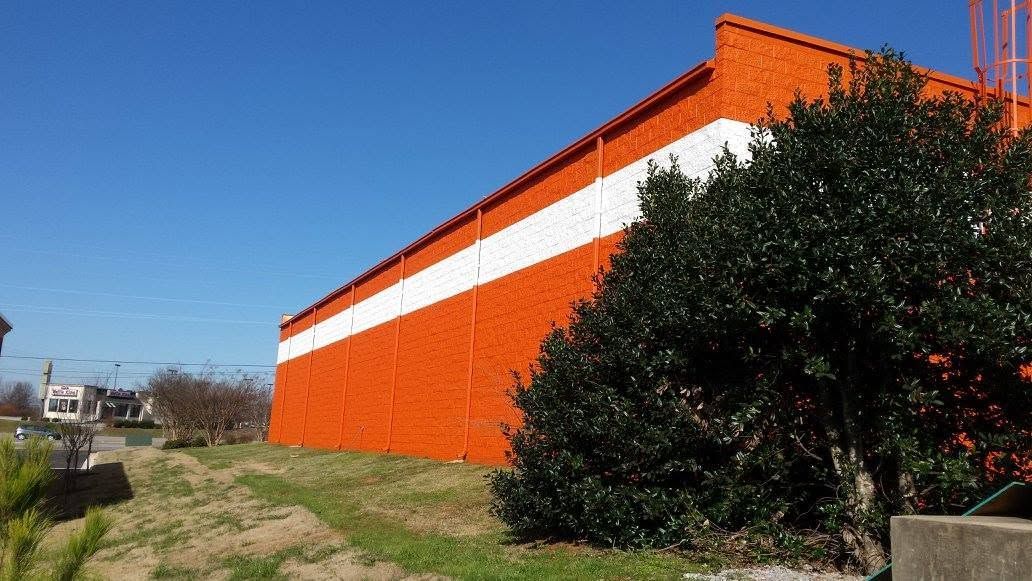 Orange building with a white stripe beside a grassy path and dense green bushes under a blue sky