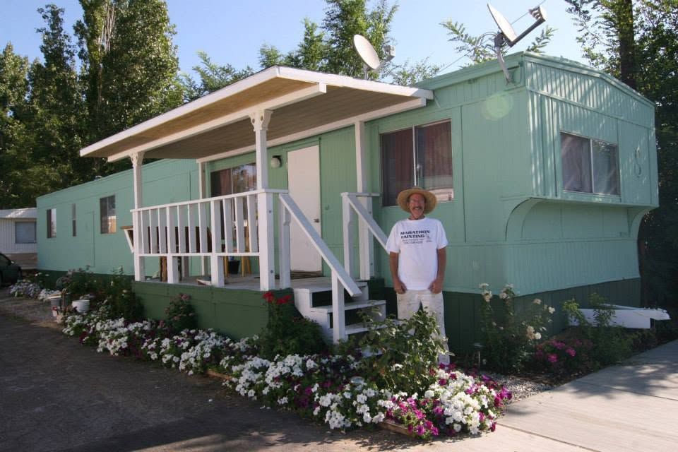 Mint-green mobile home with porch and flower beds, with a person standing outside in a hat.