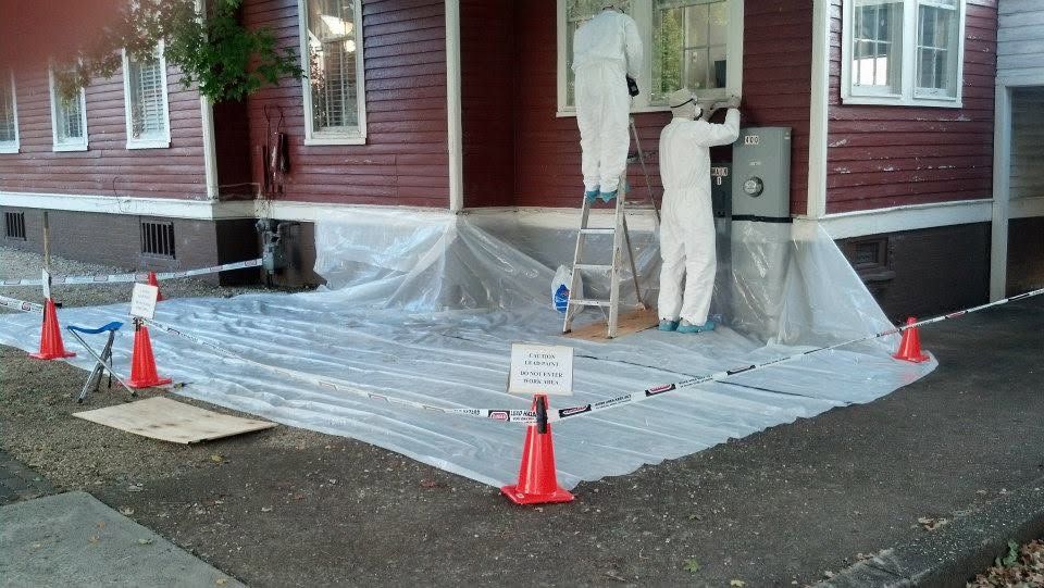 Workers in white suits spray plastic sheeting over a porch and doorway of a red house.
