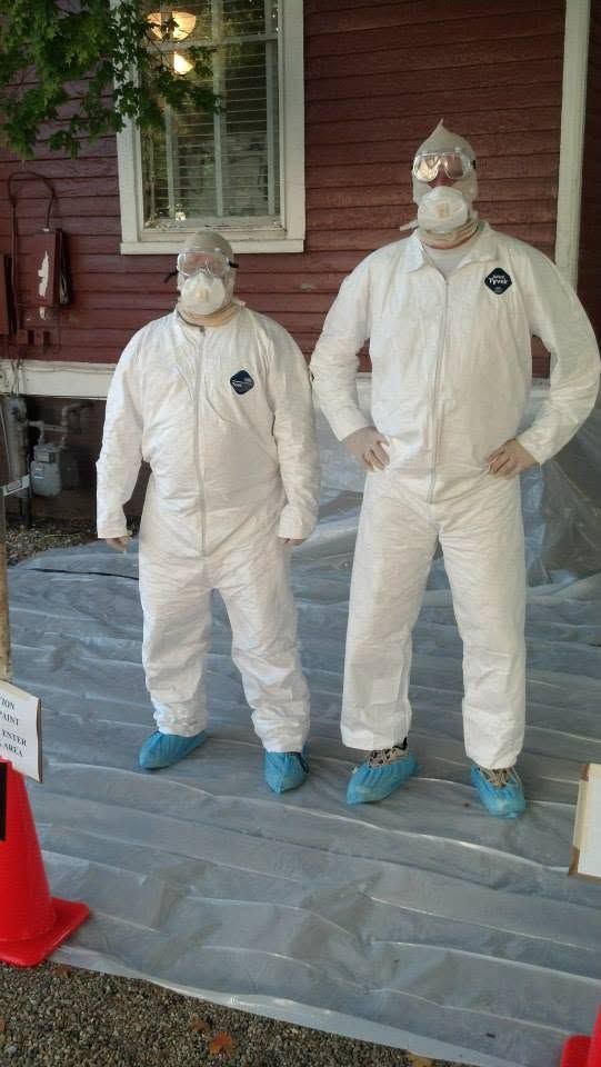 Two people in white protective suits and masks standing on a porch by a brick house.