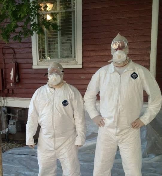Two people in white protective suits and respirators standing outside a house covered with plastic sheeting.