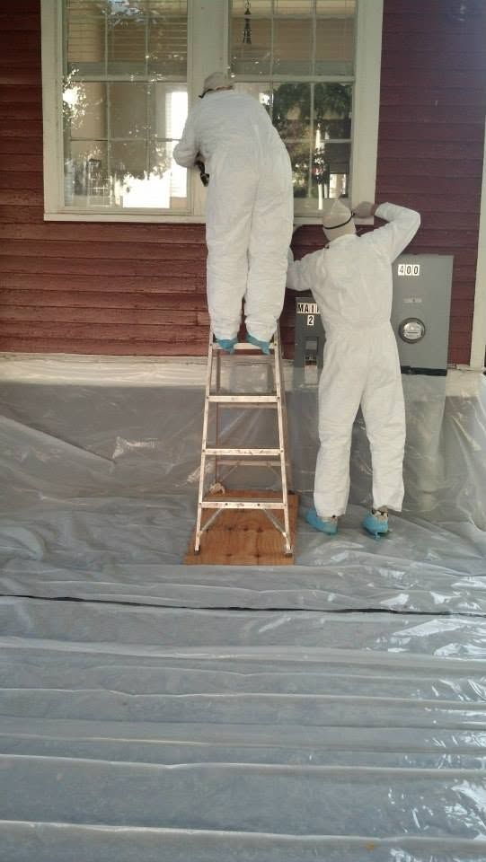 Two painters in white coveralls painting a red house exterior, one on a ladder and one beside a window.