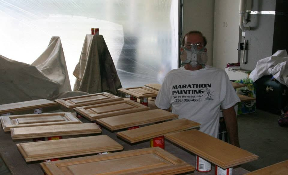 Man in a white T-shirt stands in a workshop surrounded by stacked wooden cabinet doors.