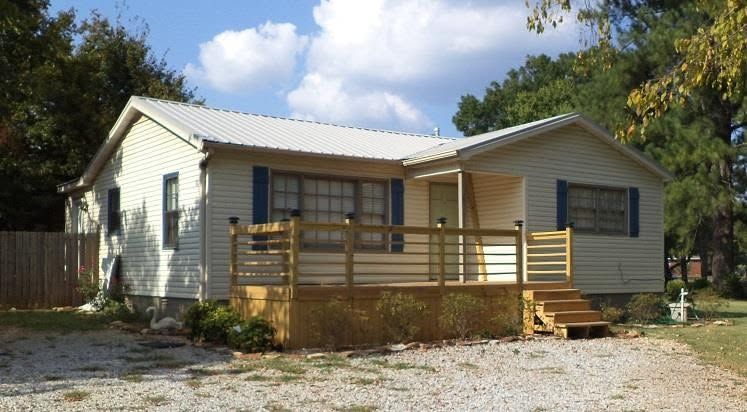 Single-story white house with a wooden front porch and steps, set on a gravel driveway under a blue sky.