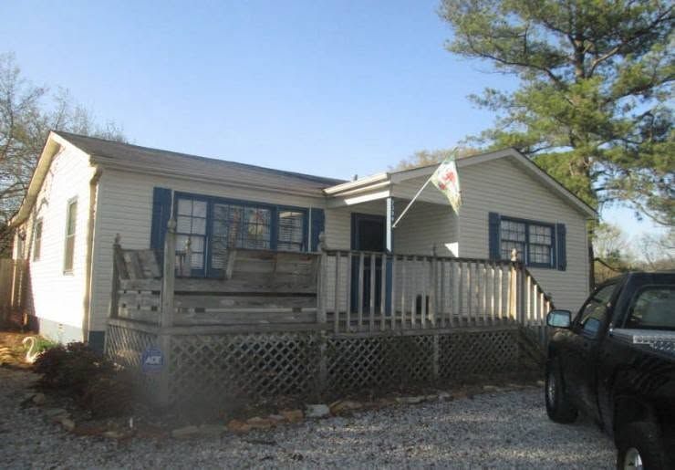 Small white house with a front porch, blue shutters, and a parked vehicle beside it under a blue sky.