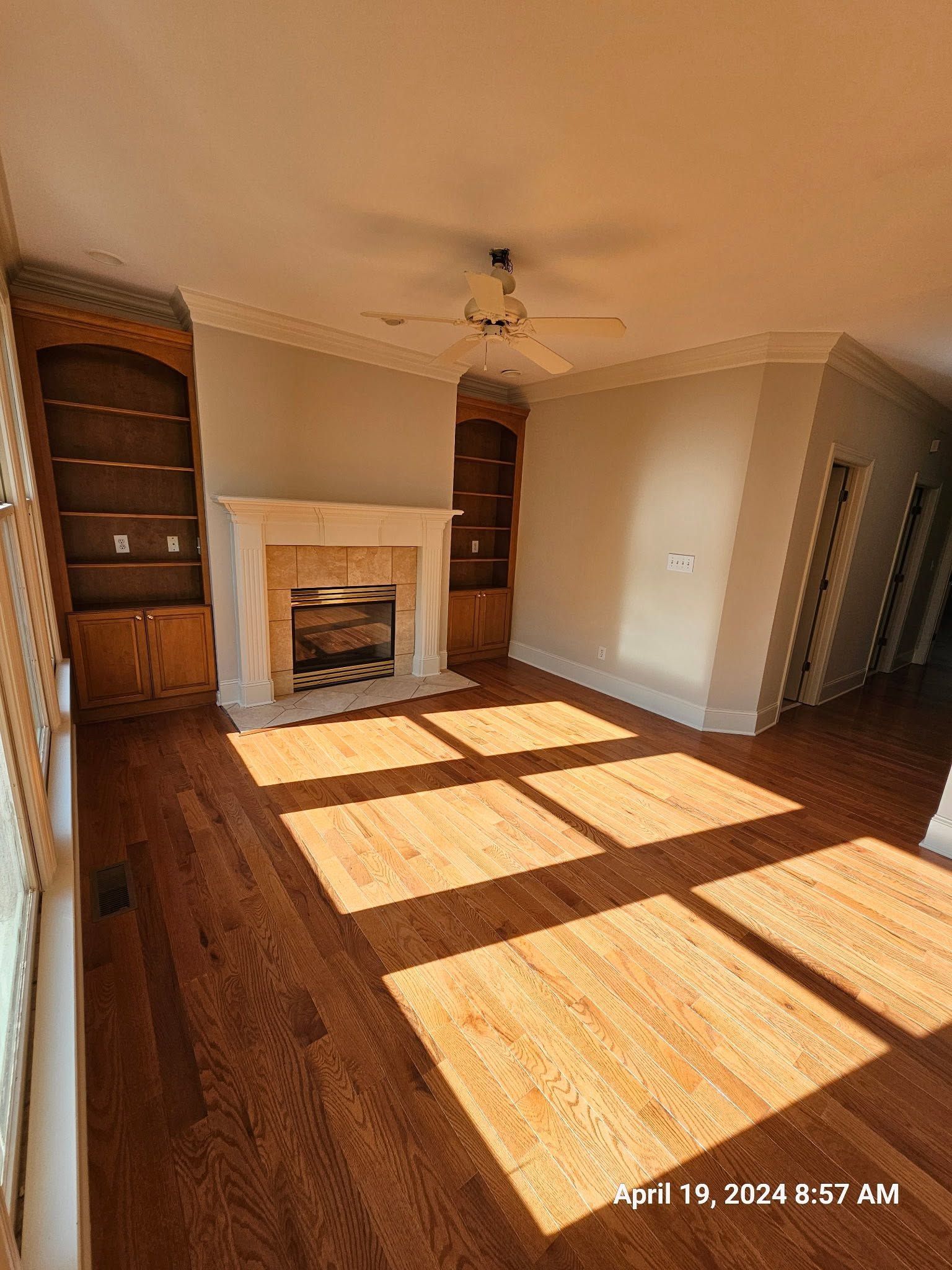 Empty living room with hardwood floors, fireplace, built-in shelves, and sunlight streaming through windows