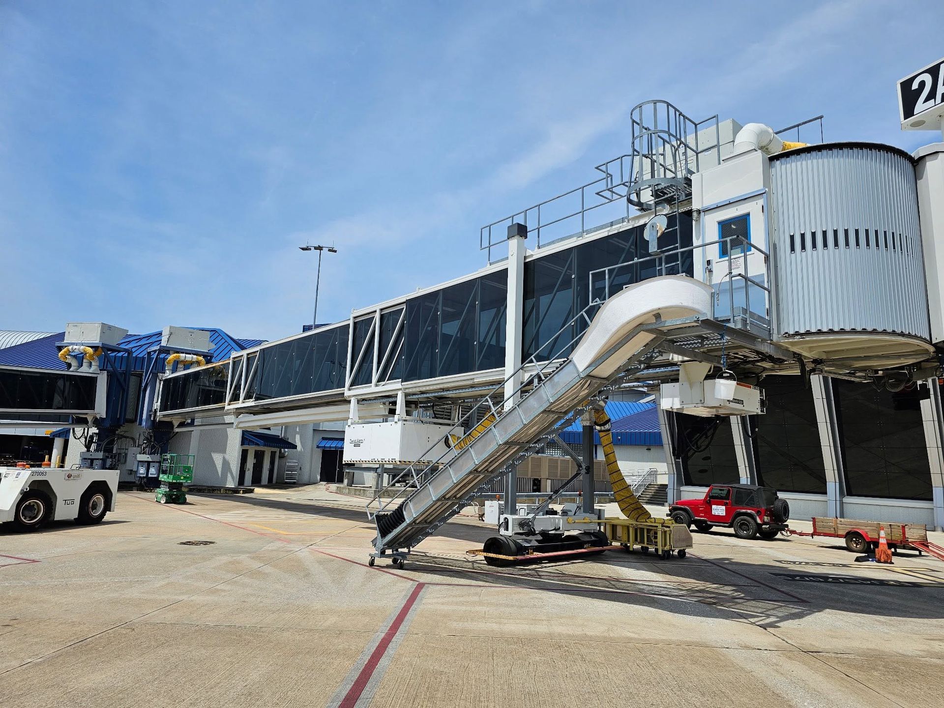 Airport terminal with a jet bridge, parked service vehicles, and a stairway on the tarmac under clear skies