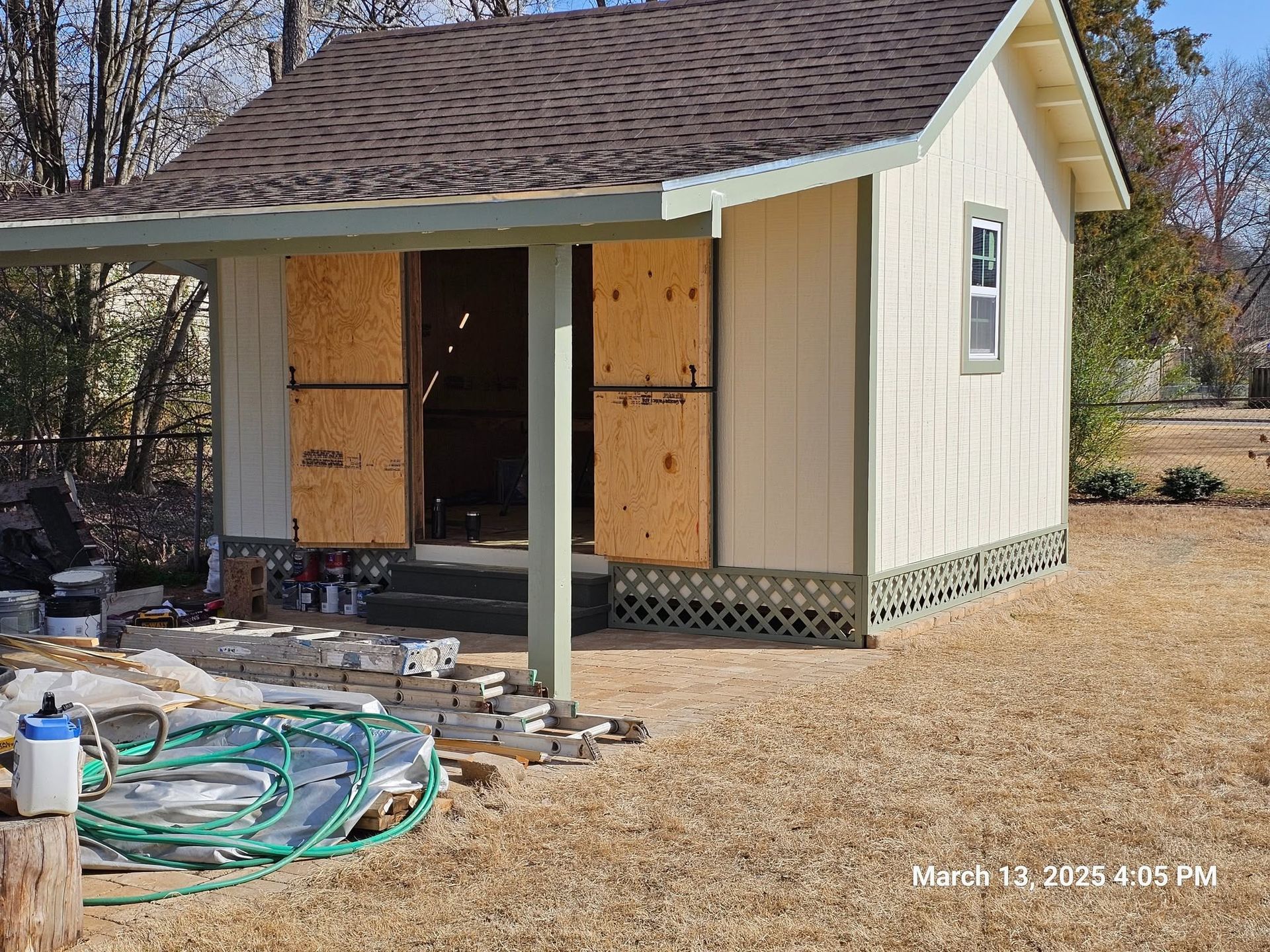 Small beige shed under construction with plywood-covered doors and a cluttered yard in front.