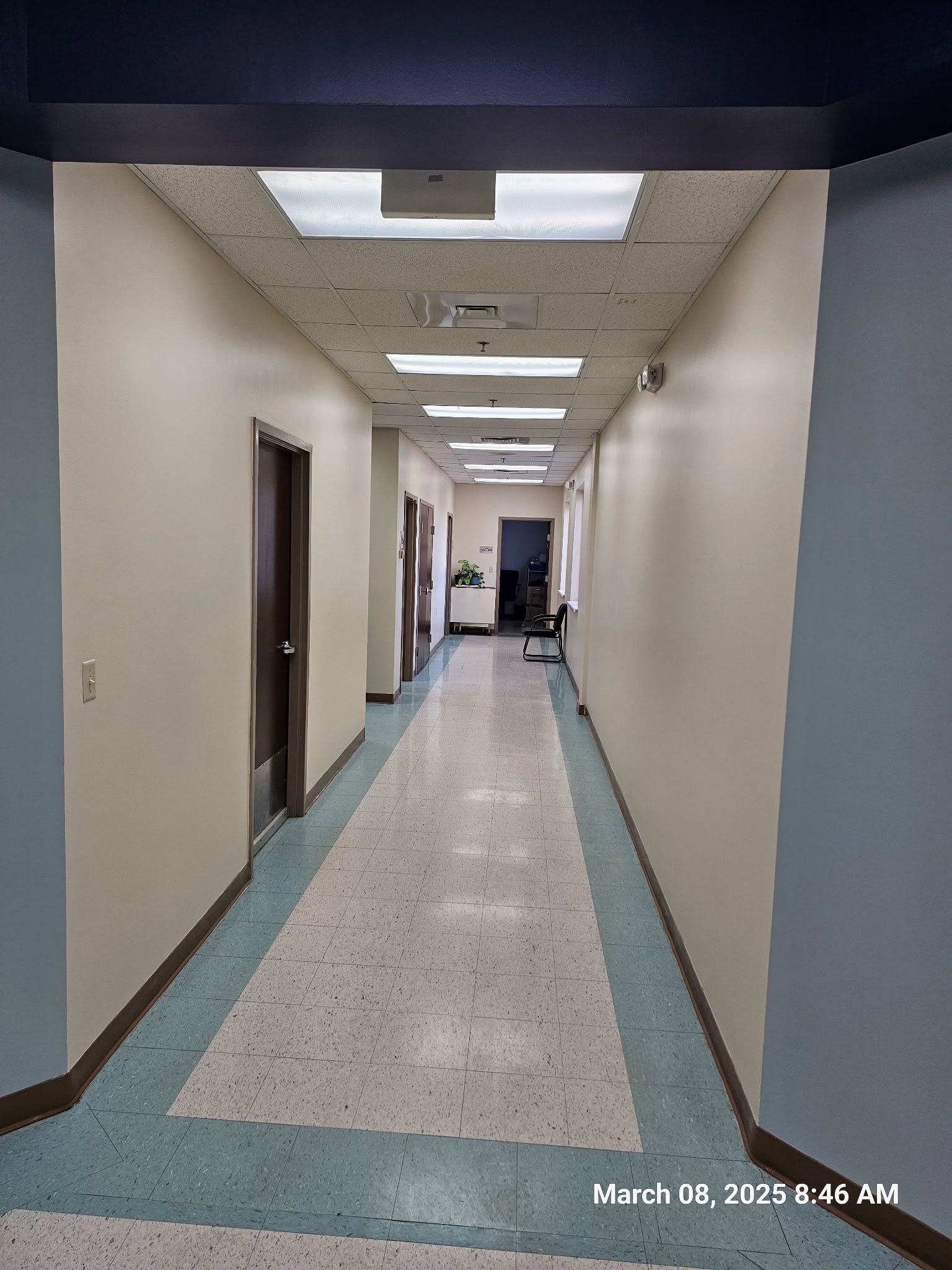 Empty hospital hallway with beige walls, blue floor trim, and fluorescent ceiling lights