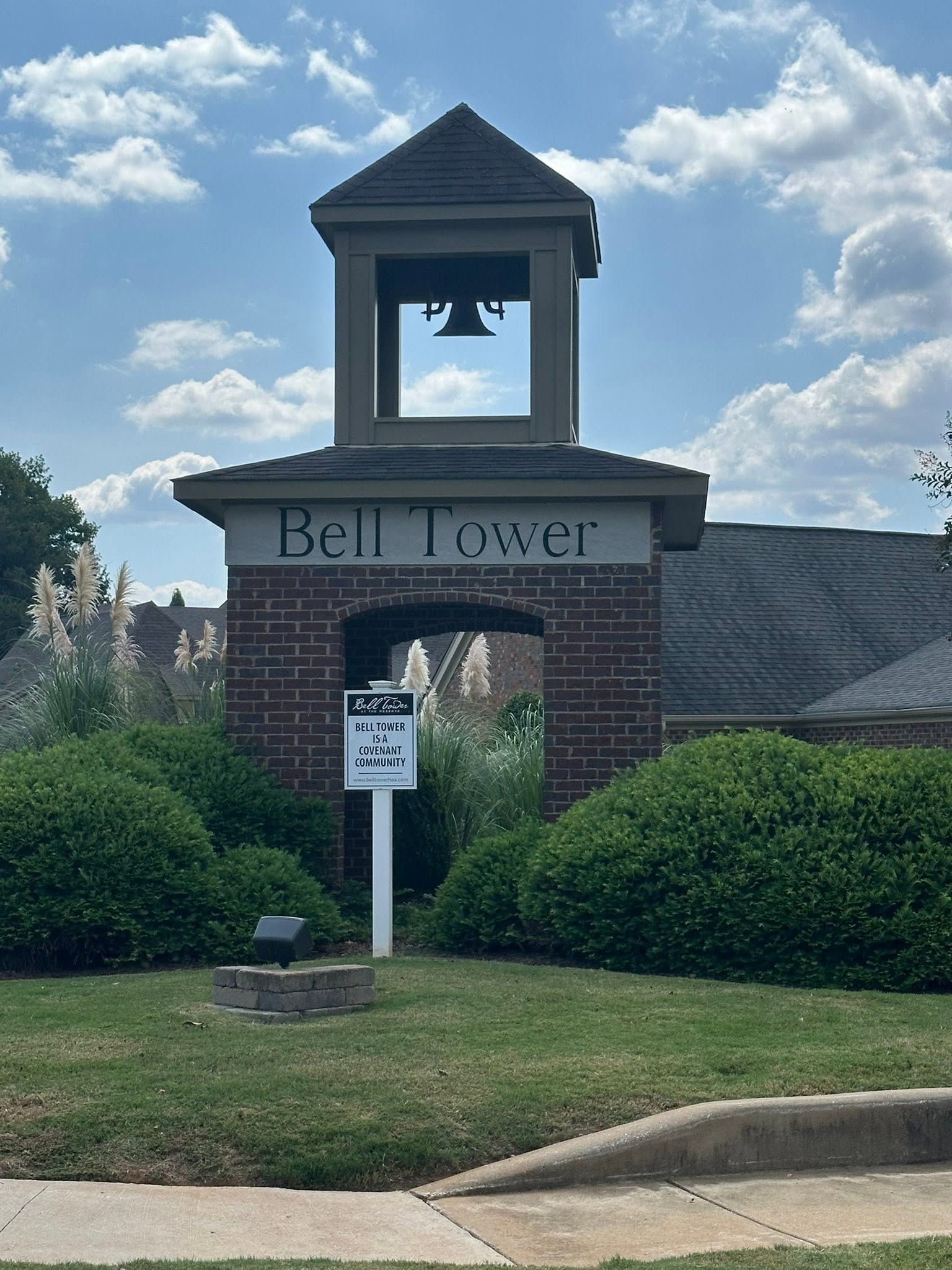 Brick bell tower entrance with arched opening and sign, set among green shrubs under a cloudy blue sky
