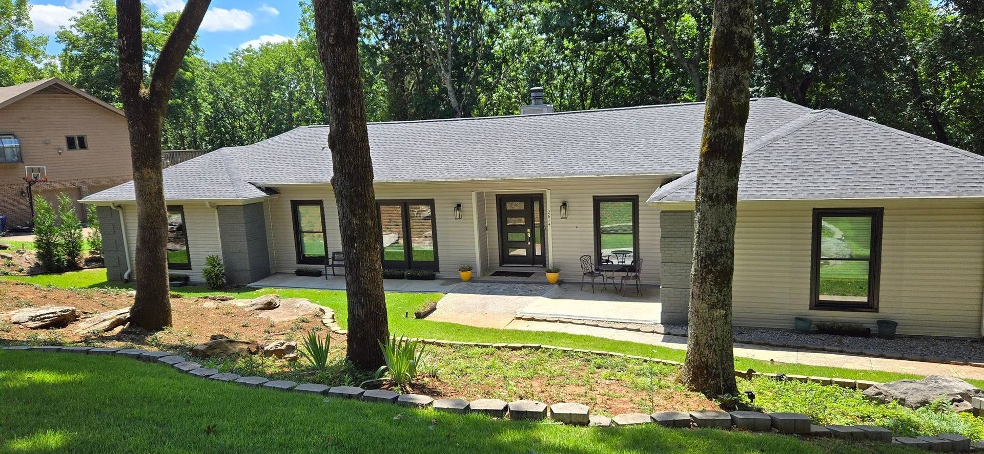 Single-story beige house with green trim and a front patio in a sunny wooded yard