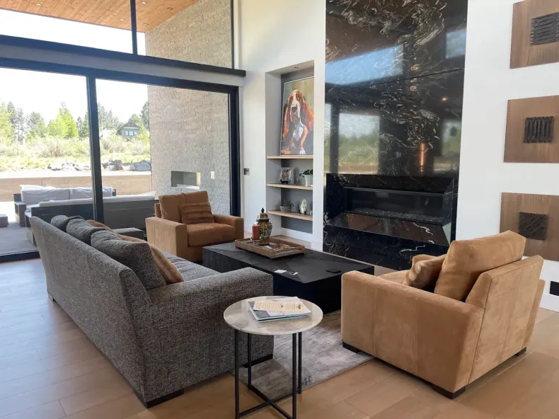 Modern living room with gray sofa, tan armchairs, fireplace, and large window overlooking trees.
