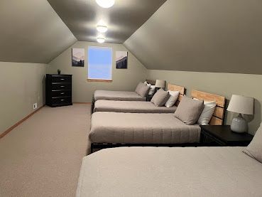 Attic bedroom with five twin beds, sloped white ceiling, dresser, bedside lamp, and small window.