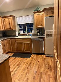 Wooden kitchen with cabinets, sink, stainless dishwasher, fridge, and window above the counter.