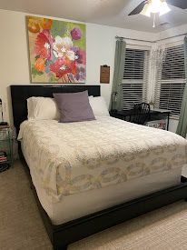 Bedroom with a neatly made bed, floral wall art, and windows with blinds.