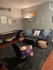 Cozy living room with gray sofa, black rug, piano, wall art, and woven stools