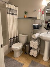 Small bathroom with white toilet, pedestal sink, shelves of rolled towels, and a shower curtain