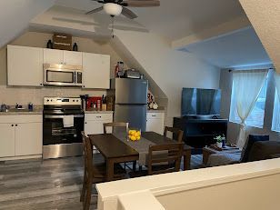 Open loft kitchen and living area with dining table, TV, sofa, and large windows.