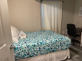 Small bedroom with a bed, blue floral bedspread, window curtain, and desk chair.