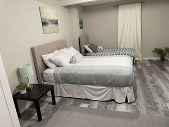 Bright bedroom with two beds, gray rug, nightstand, lamp, and curtained window.