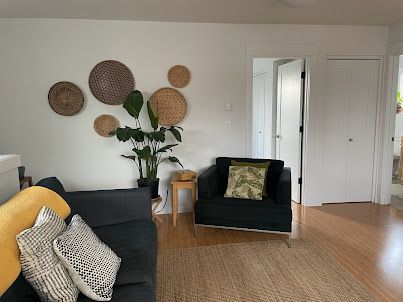 Modern living room with dark sofas, woven wall decor, a large plant, and open white doors.