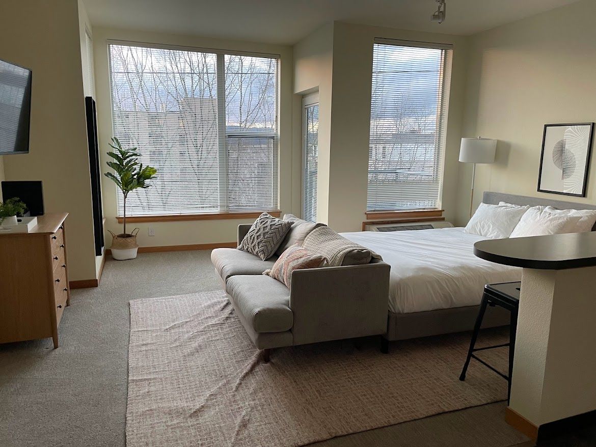 Bright bedroom with a bed, sofa, desk, and large windows with blinds