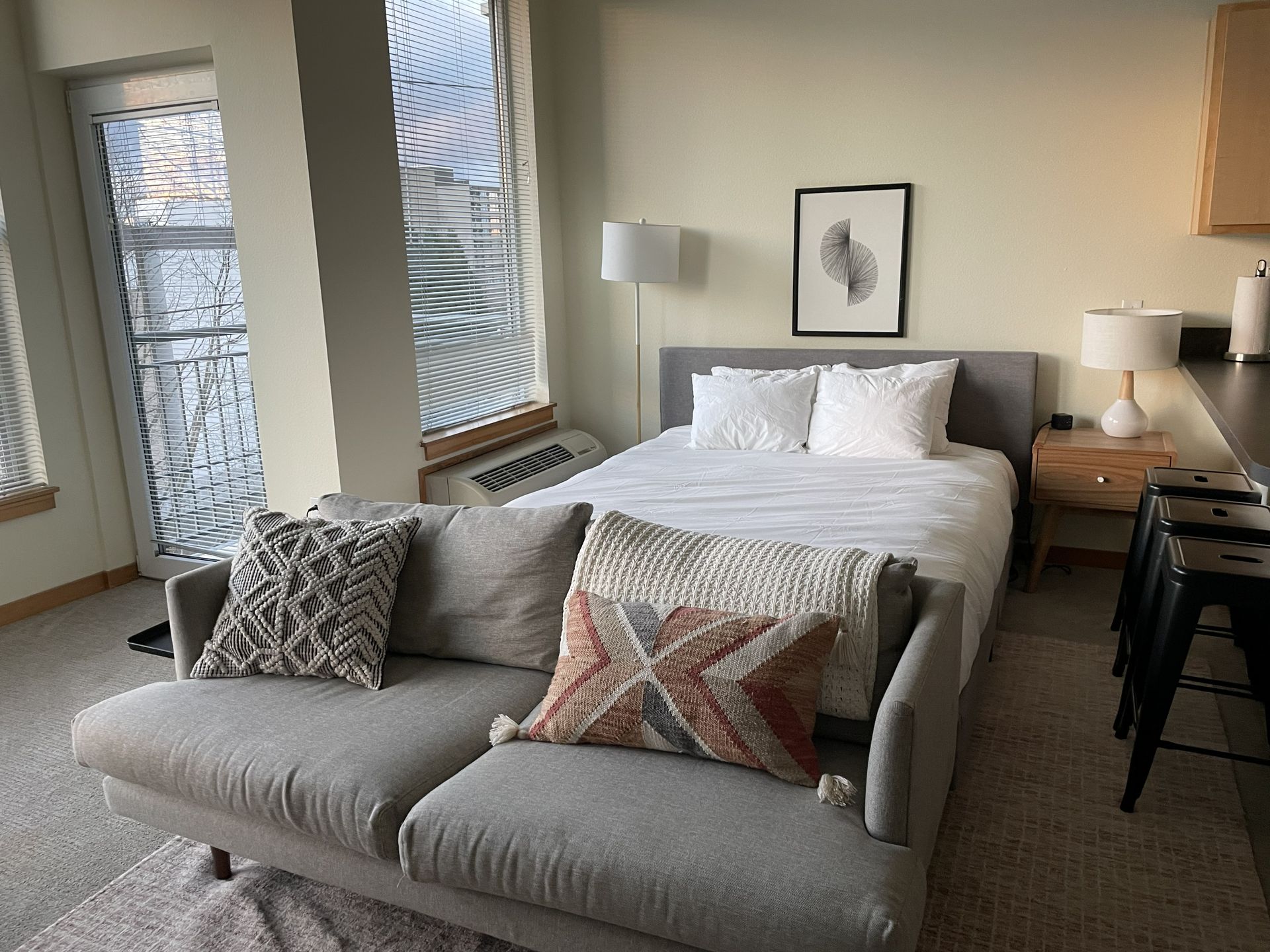 Cozy bedroom with a bed, chaise lounge, nightstands, lamp, and patterned pillows near large windows