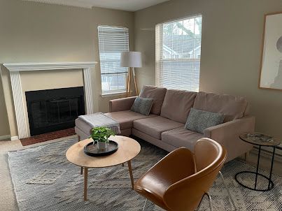 Living room with beige sectional, tan chair, round coffee table, fireplace, and windows with blinds