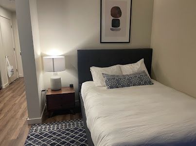 Small bedroom with a neatly made bed, bedside lamp, framed art, and patterned rug.