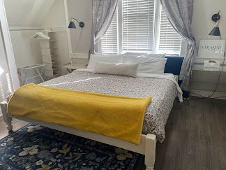 Bright bedroom with a white bed, yellow throw, patterned rug, and curtains over a window.