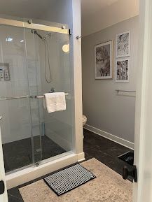 Modern bathroom with glass shower stall, beige rug, dark floor, and framed wall art.