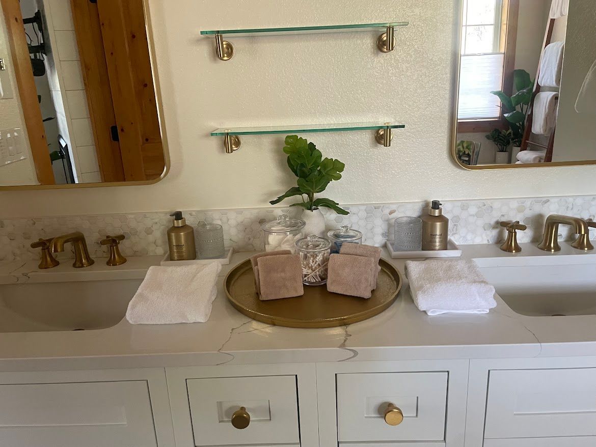 Bathroom vanity with a centered plant, gold tray, candles, towels, and brass fixtures beneath a mirror.