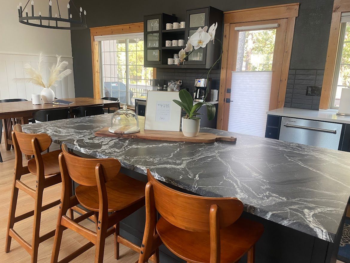 Modern kitchen with a large dark marble island, wooden bar stools, and bright windows.