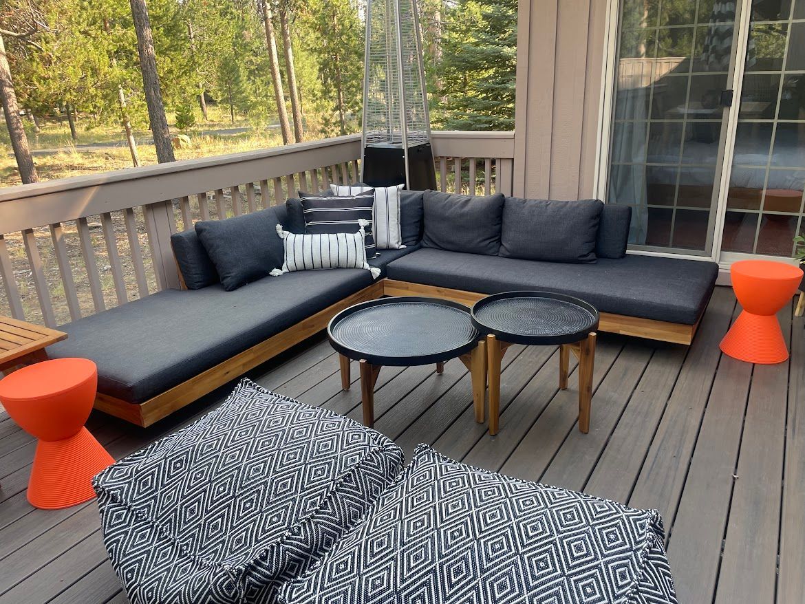 Wooden deck with gray sectional sofa, round tables, patterned lounge chairs, and orange stools overlooking trees