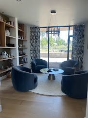 Modern living room with blue chairs around a round table, bookshelf, and large window with curtains.