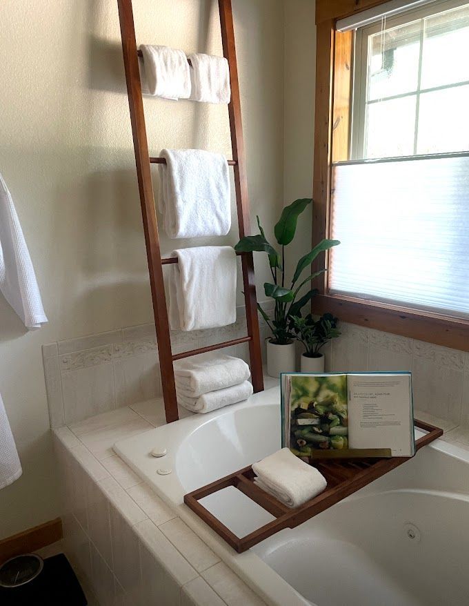 Bathroom with white tub, wooden towel ladder, plant, and framed decor by a window