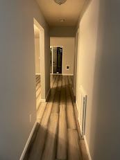 Narrow hallway with wood floors, beige walls, and a doorway at the end.