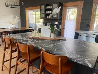 Modern kitchen with large black marble island, wooden chairs, and sunlight through windows