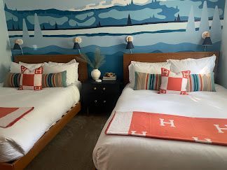 Twin beds in a cozy room with blue mountain mural, white bedding, and coral accent blankets.