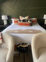 Modern bedroom with a neatly made bed, striped pillows, bedside lamps, and two white chairs in the foreground