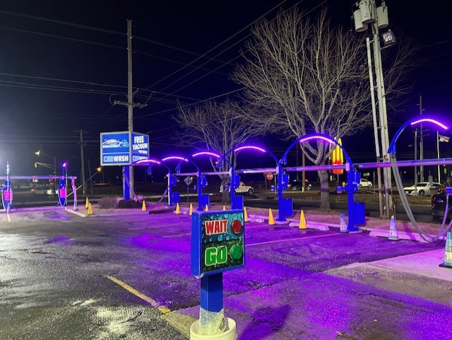 An illuminated car wash entrance at night with purple neon light arches, traffic cones, and a