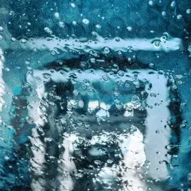 Raindrops on a glass surface create a blurred, distorted view of a bright interior light against a blue-toned background.